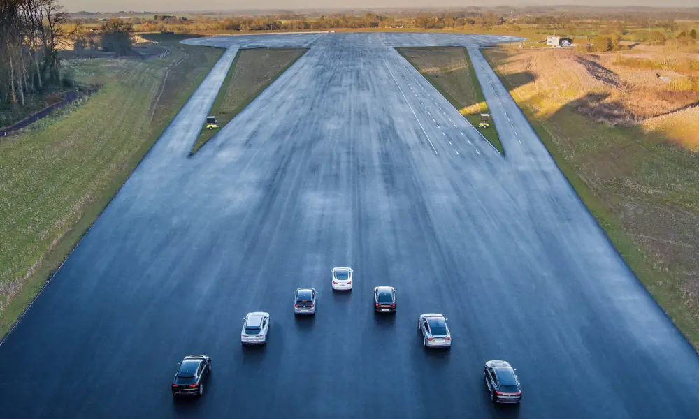 A drone shot of seven cars in a V-formation on a very wide tarmacked strip at a multi lane vehicle testing site.