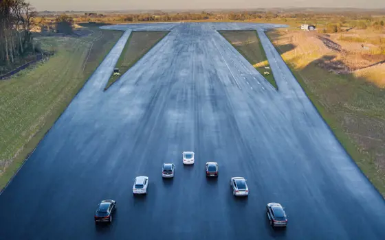 A drone shot of seven cars in a V-formation on a very wide tarmacked strip at a multi lane vehicle testing site.