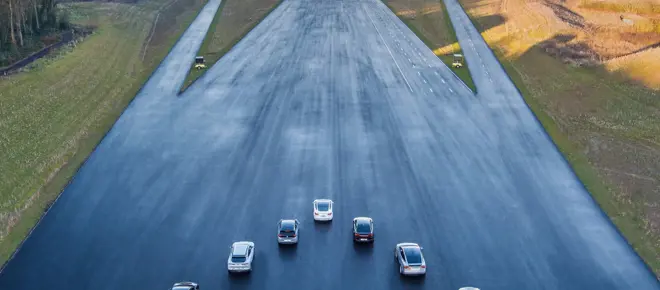 A drone shot of seven cars in a V-formation on a very wide tarmacked strip at a multi lane vehicle testing site.