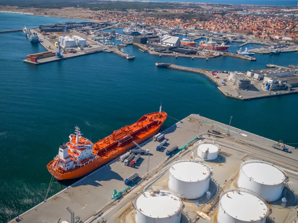 A ship being refueled at a bunkering facility, with trucks carrying fuel and enormous white storage tanks visible on the dock.