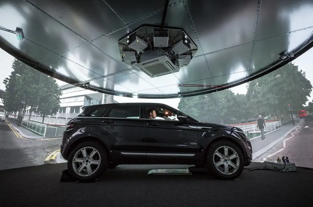 A person inside a car indoors with a virtual screen surrounding them that has scenarios being played out on a driving simulator.