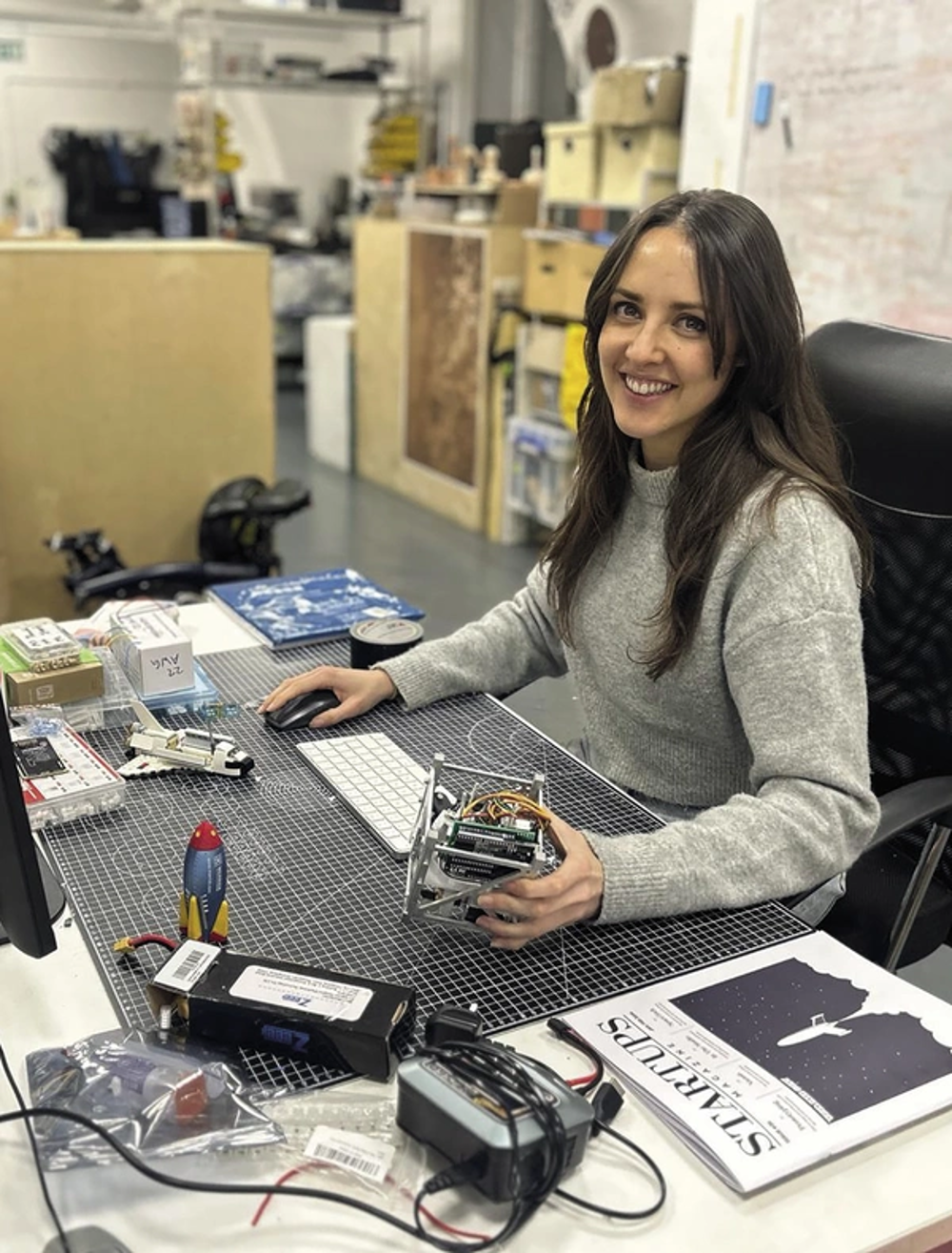 A woman with brown hair sitting at a desk smiling, holding an electronic prototype.