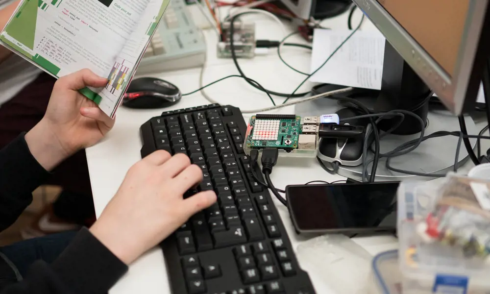 A child holding a book and typing on a keyboard, with a Raspberry Pi connected to the computer in front of them.