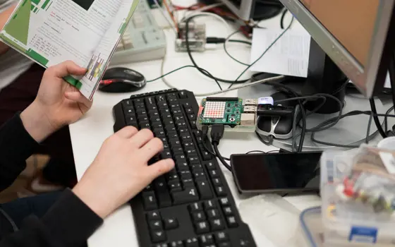 A child holding a book and typing on a keyboard, with a Raspberry Pi connected to the computer in front of them.
