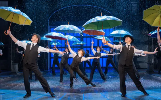 Dancers on a stage performing in the "singin' in the rain" musical, pointing umbrellas towards the ceiling. Water is falling down on them and they are splashing in puddles of water as they dance. 
