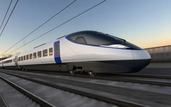 Artist’s impression of a sleek, modern high-speed train traveling on railway tracks. The train has a streamlined design with a pointed nose, white body, and blue accents around the doors. Overhead electric wires run above the tracks, and the background shows a blurred landscape under a clear sky at sunset, suggesting speed and motion.