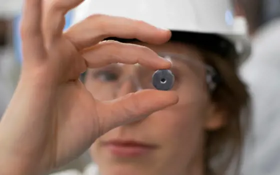 A close-up of a scientist wearing goggles and a safety hat holding a small metal round object. 