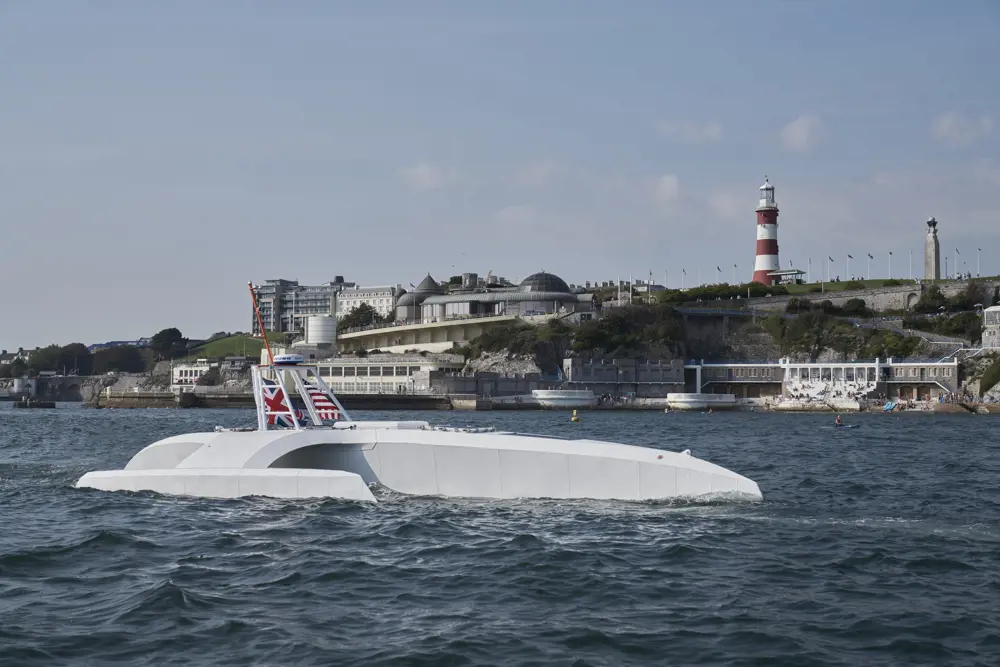 The top of the Mayflower submarine above water with the UK flag attached to it and a town in the background. 