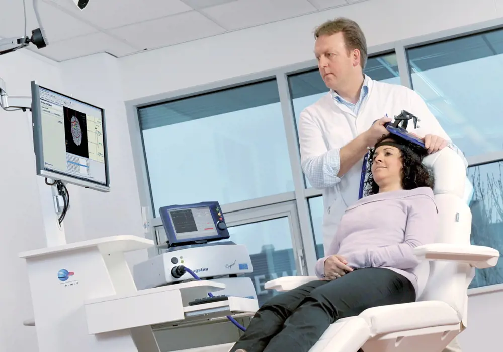 A person using a magnetic stimulator coil device on another person's head who is sitting in a chair.