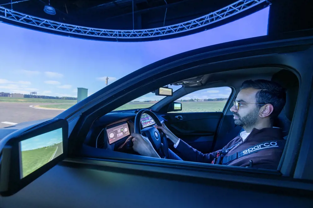 A male engineer wearing glasses, sitting in the driving seat of a simulator with a wrap around screen.