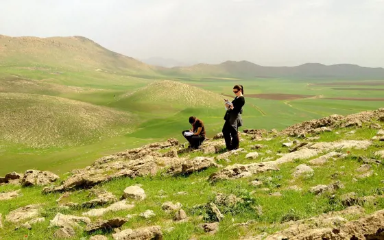 Two people stand on the side of a hill writing on clipboards with other hills in the background