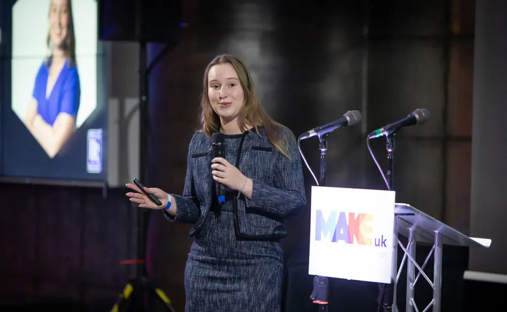 Person presenting on stage with a microphone at an event, with a lectern and presentation screen in the background.