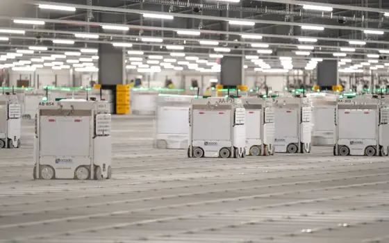 A well lit large room at the Erith warehouse, containing cube-shaped robots with wheels, on a large grid as a floor. 