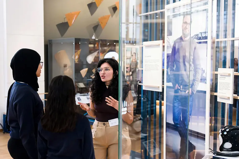 Person speaking to two students inside a museum gallery, with display cases and exhibits visible through glass panels.