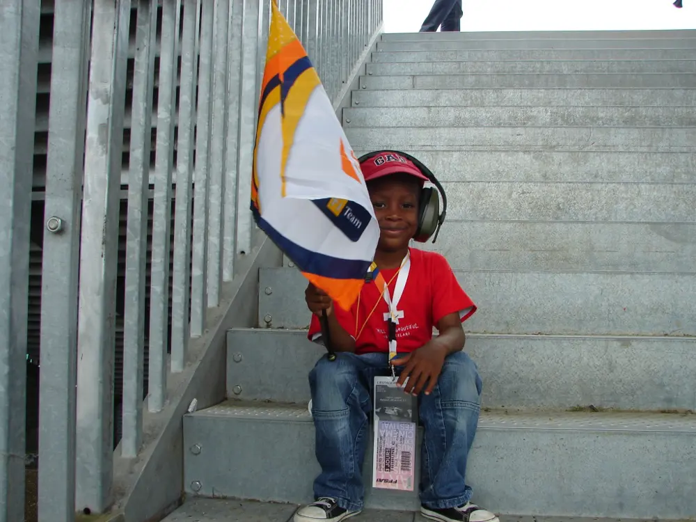 A young boy sitting on the steps up to a spectator stand, wearing ear defenders and holding a small flag.