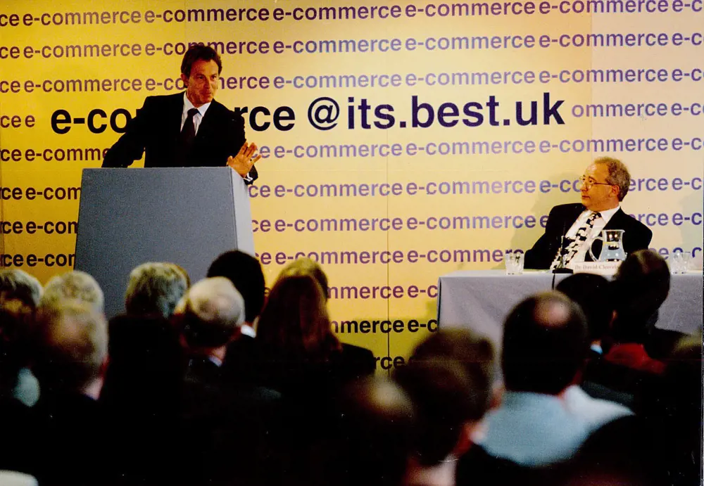 Tony Blair standing at a podium next to David Cleevely FREng sitting at a desk on stage. 