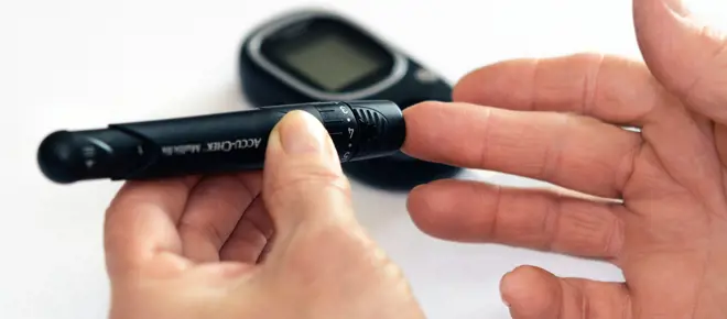 A pen-shaped device taking a pinprick of blood from someone's middle finger with a handheld sensor screen in the background.