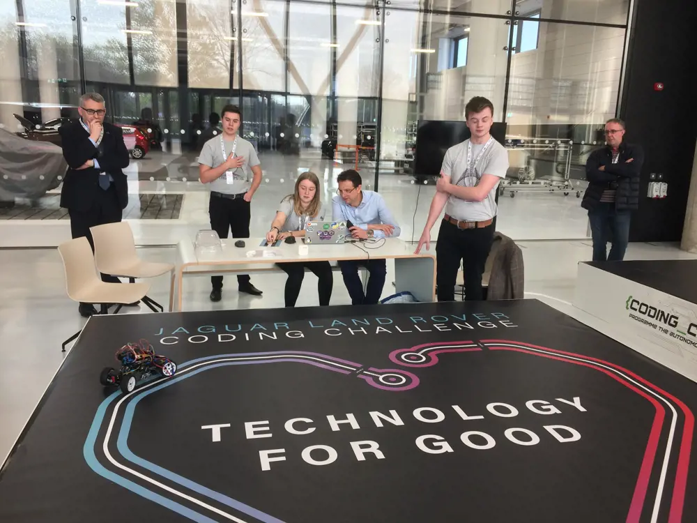 Nick Rogers sitting at a desk next to younger engineers who have entered into the Jaguar Land Rover coding challenge with a small model vehicle on the floor in front of the desk.