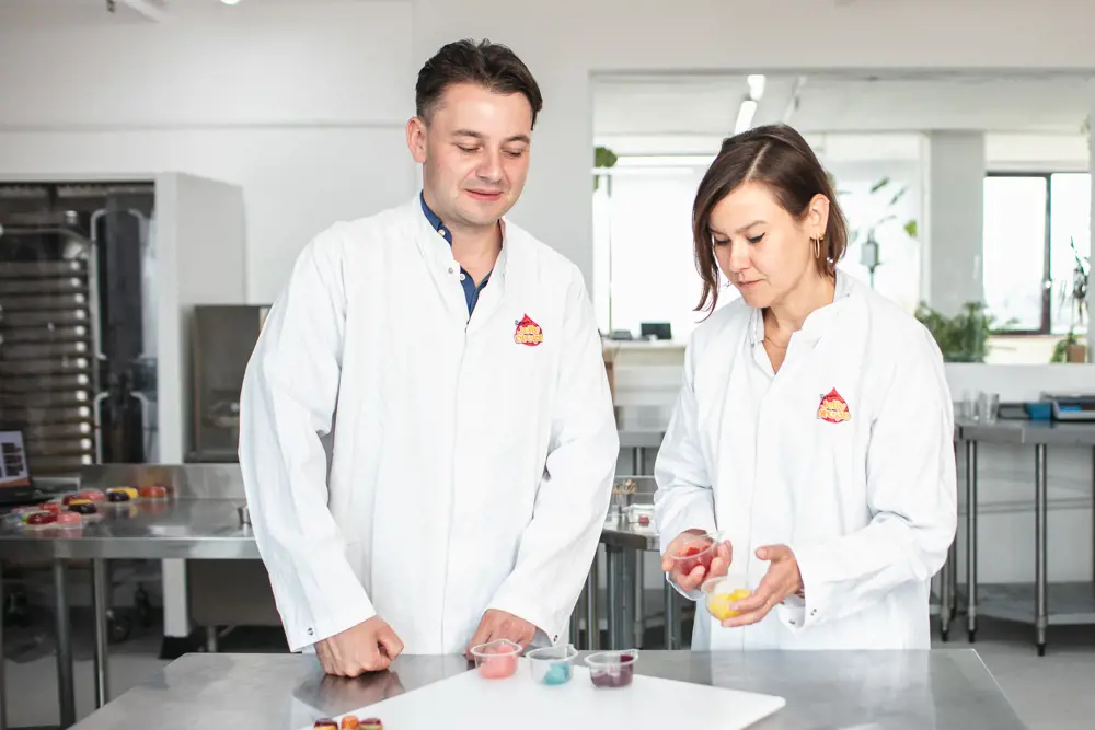 Two people standing in a test kitchen, wearing chef whites-style lab coats and holding pots of brightly coloured sweets