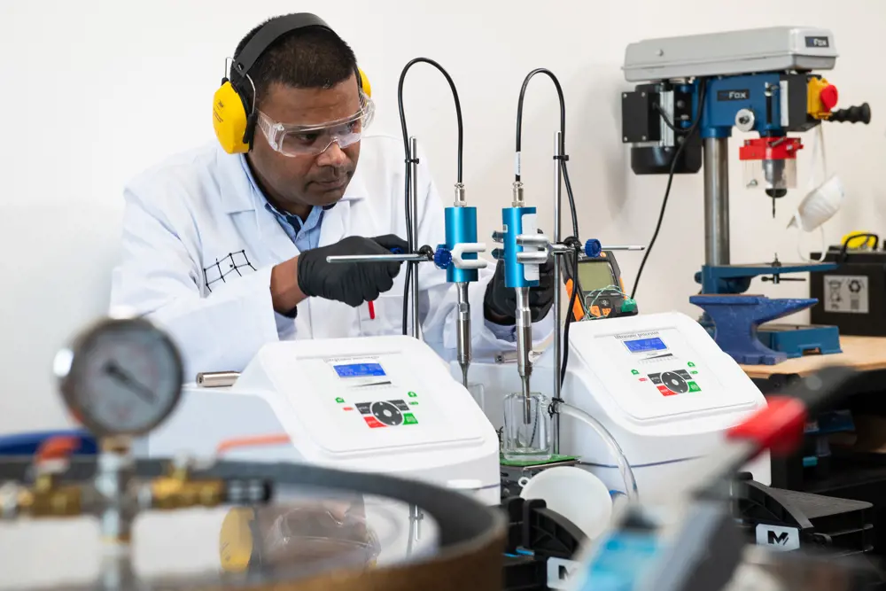 A man with a lab coat and yellow ear defenders focuses on adjusting his setup on a lab bench.