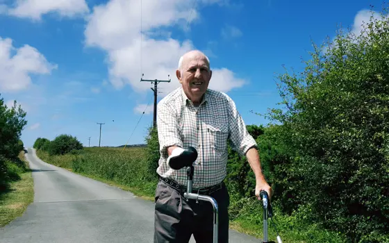 Eilian walking on a road lined with green hedges on a sunny day. He is using his adapted three-wheeled rollator.