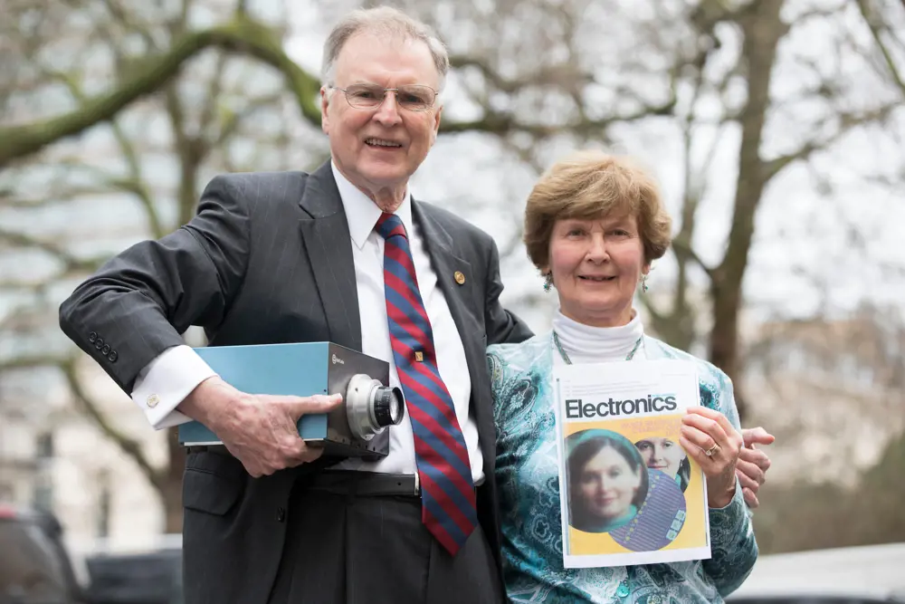 Micheal Tompsett holding a colour camera, with his arm around his wife, Margaret, who holds a photo of herself printed on the cover of an electronics magazine. 