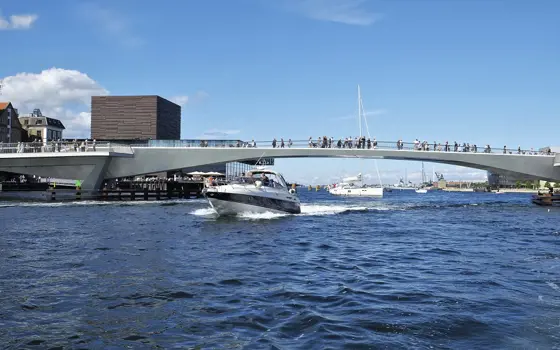 A boat driving under the Inner Harbour Bridge in Copenhagen. People are walking across the bridge. 