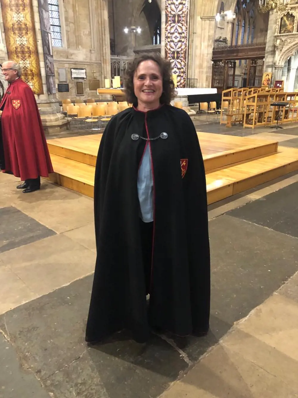 Penny has short dark hair, she is smiling in a cathedral, wearing a black robe.