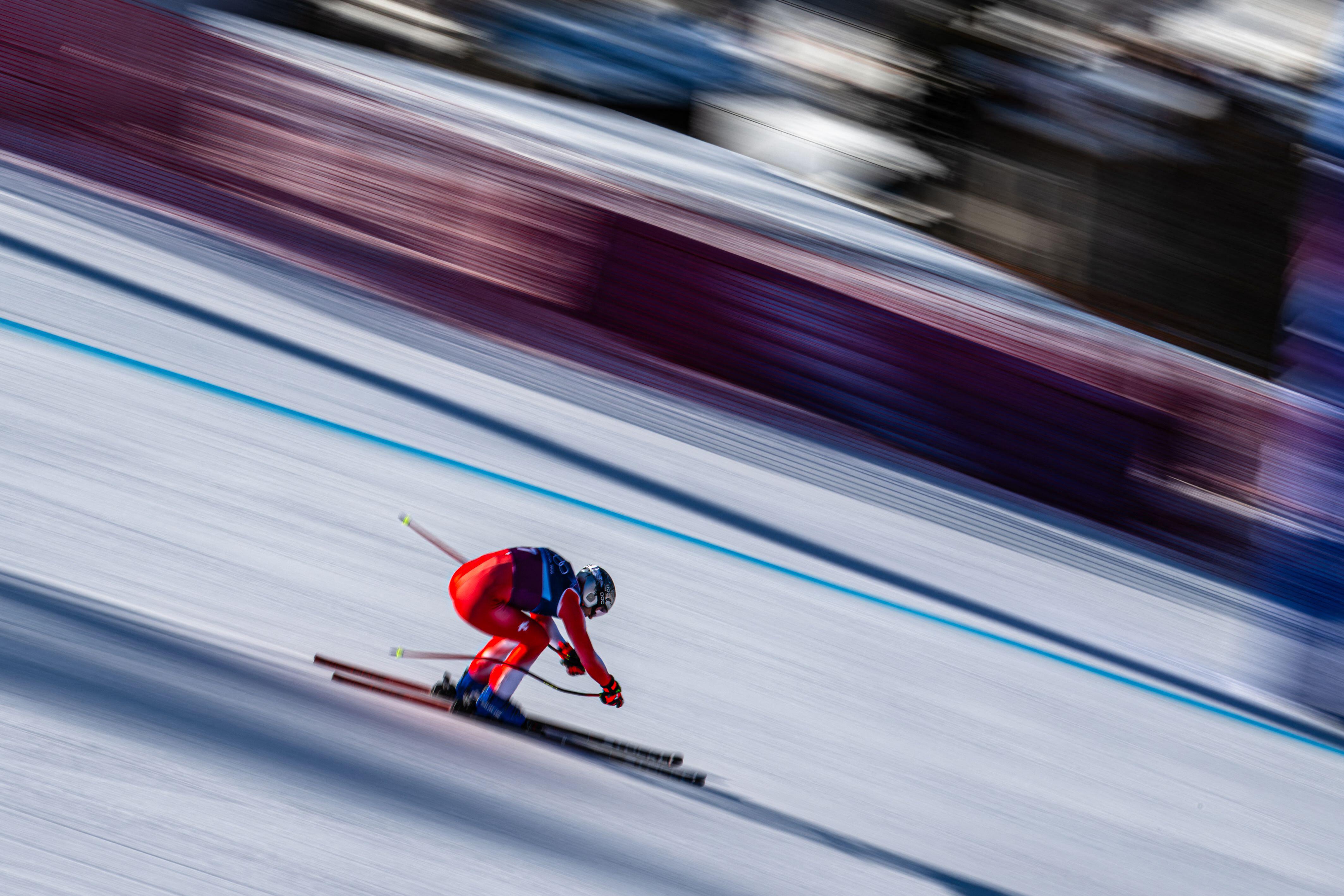 Switzerland's Marco Odermatt, wearing a red racing ski suit leans forward into his skis at high speed on a snowy slope, with motion blur showing rapid movement.