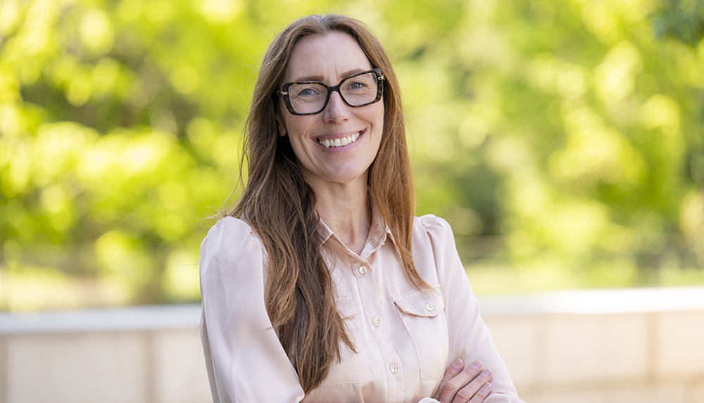 A woman with long dark hair and wearing glasses stands in front of a bush outside while smiling at the camera