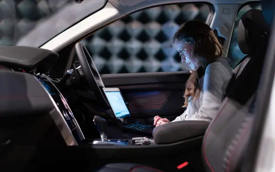 A female electronics engineer runs tests on a laptop while sitting in the driver's seat of a car.