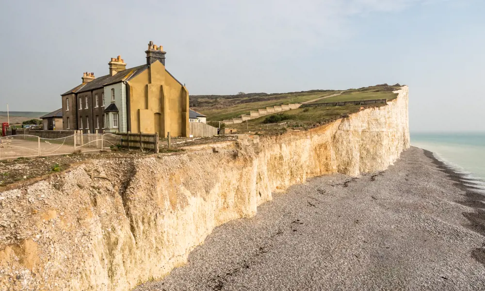 Four houses pictured on a cliff edge, with a pebble beach below it. 