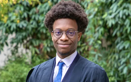 A man with an afro and glasses smiles for a photo, wearing a suit and matriculation gown to enrol at the University of Cambridge.