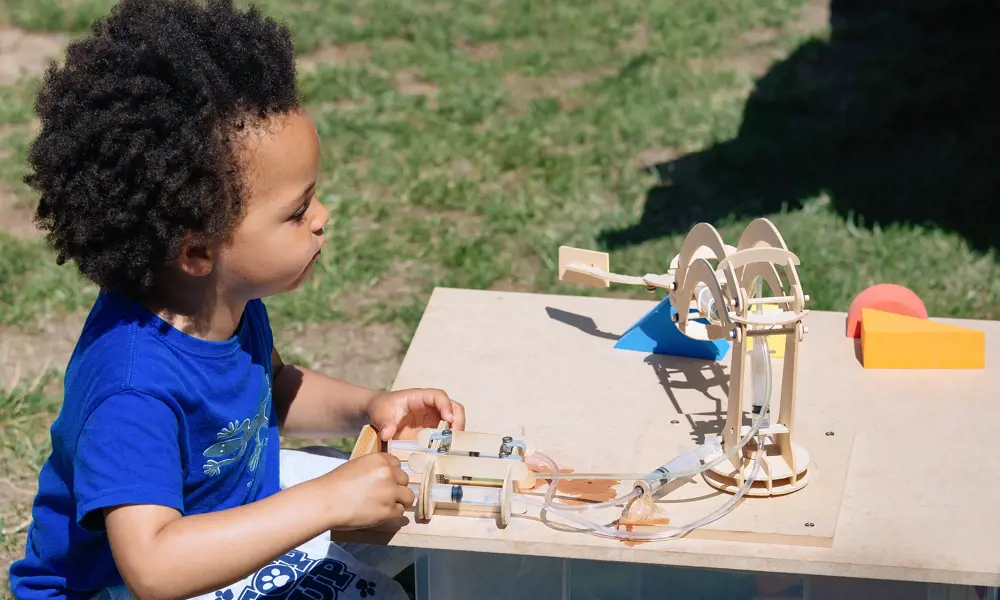 A young child playing outside with a wooden model containing syringes connected to plastic tubes. 
