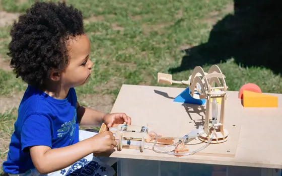 A young child playing outside with a wooden model containing syringes connected to plastic tubes. 