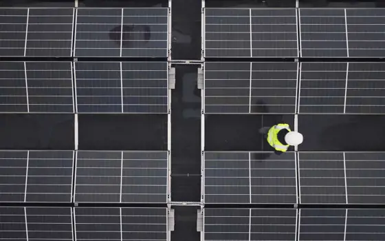 An aerial view of solar panels on a roof with an apprentice engineer in a high visibility jacket and helmet inspecting them.