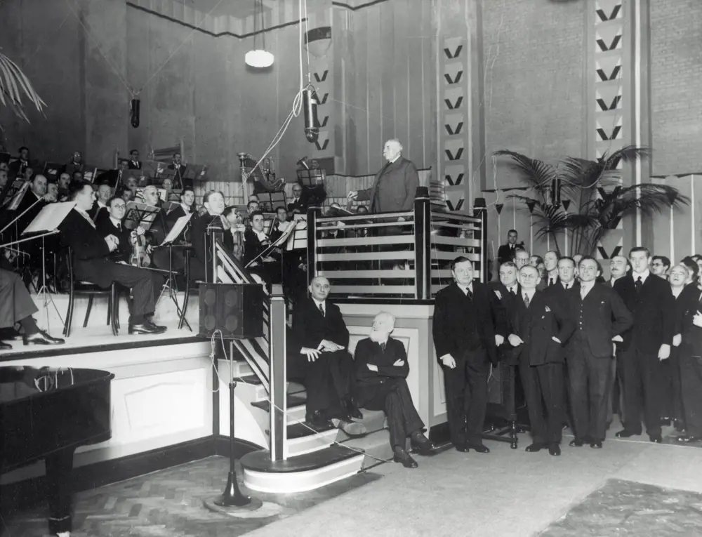 A greyscale image of an orchestra being conducted by Sir Edward Elgar, with a group of people standing near to listen to the acoustics. 