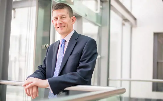 A headshot of Dr Shaun Fitzgeralsn FREng wearing a suit, smiling and resting his hands on a banister.