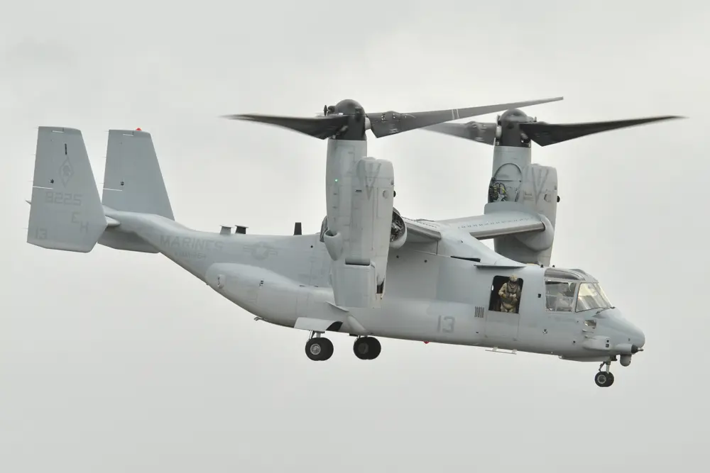 A V-22 Osprey VTOL aircraft in the air, with a marine standing looking out of its side hatch.