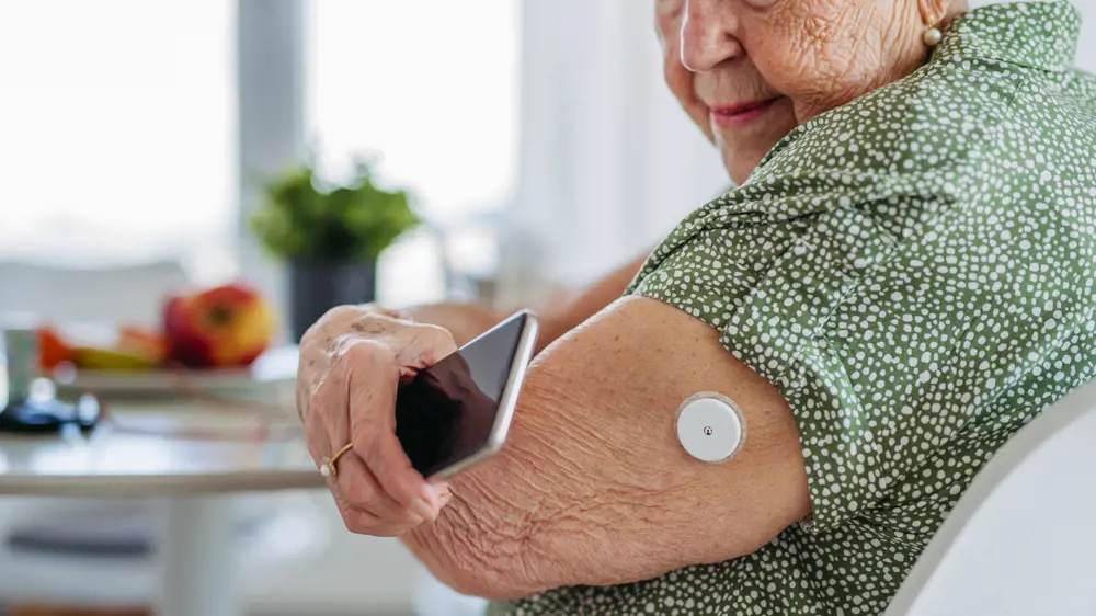An older white woman wearing a green blouse holds her phone up to her arm, on which she is wearing a continuous glucose monitoring device