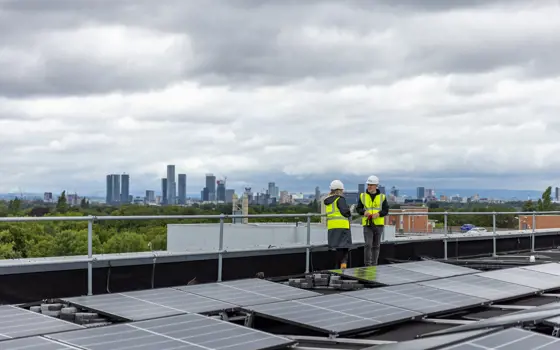 Two engineer colleagues speaking to each other on top of a building with solar panels, with a skyline in the background.