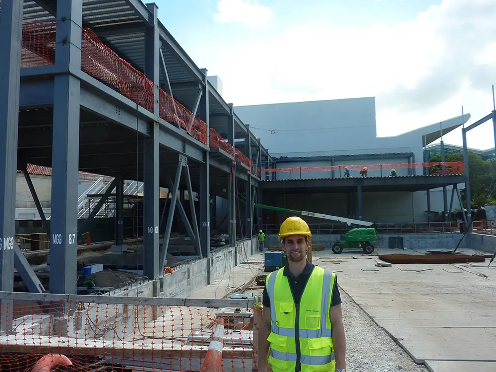 Steel-framed building under construction with safety netting, machinery, and a worker in a high‑visibility vest and hard hat in the foreground.