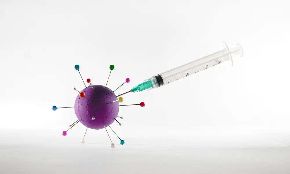 A syringe with the needle sticking into a purple sphere with long pins sticking out of it, which is a model of a covid virus particle