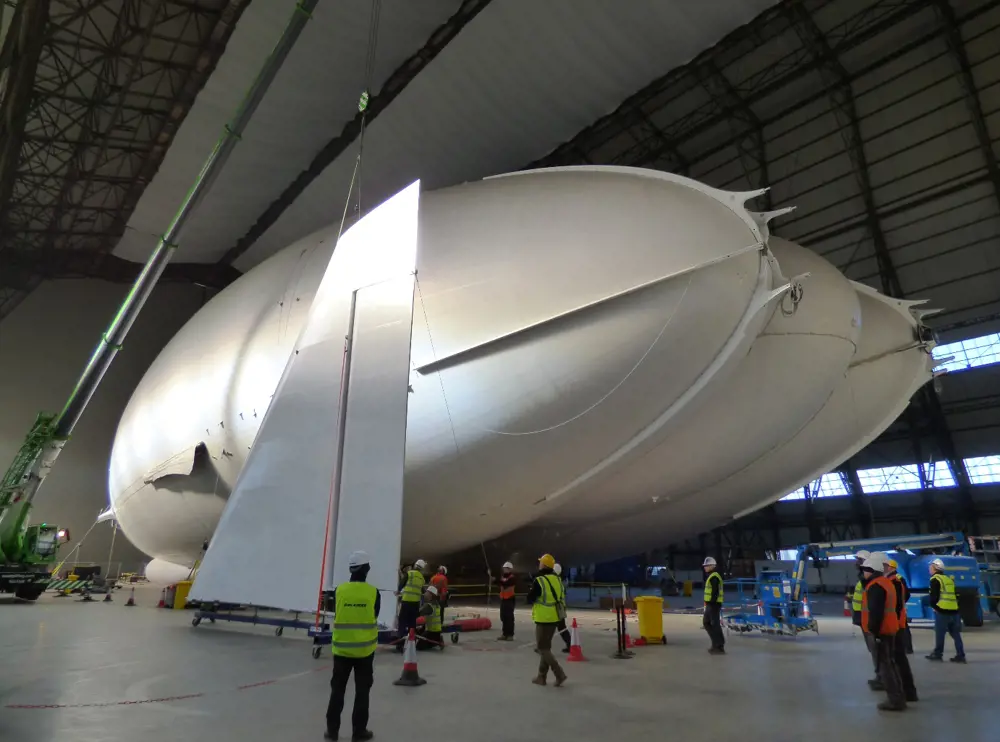 A group of people in safety gear in a factory standing under the Airlander.