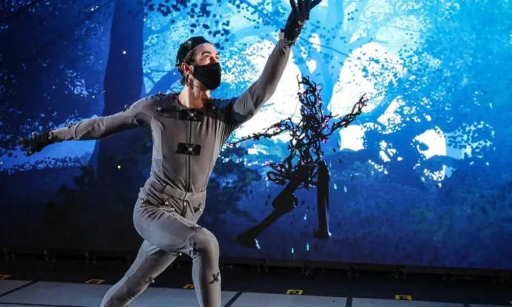 Someone in a motion capture suit, who is controlling the motion of a woodland avatar on a digital animation in the background.