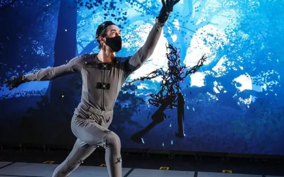 Someone in a motion capture suit, who is controlling the motion of a woodland avatar on a digital animation in the background.