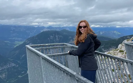 A woman with red hair wearing sunglasses stands on a lookout, looking over a mountain range