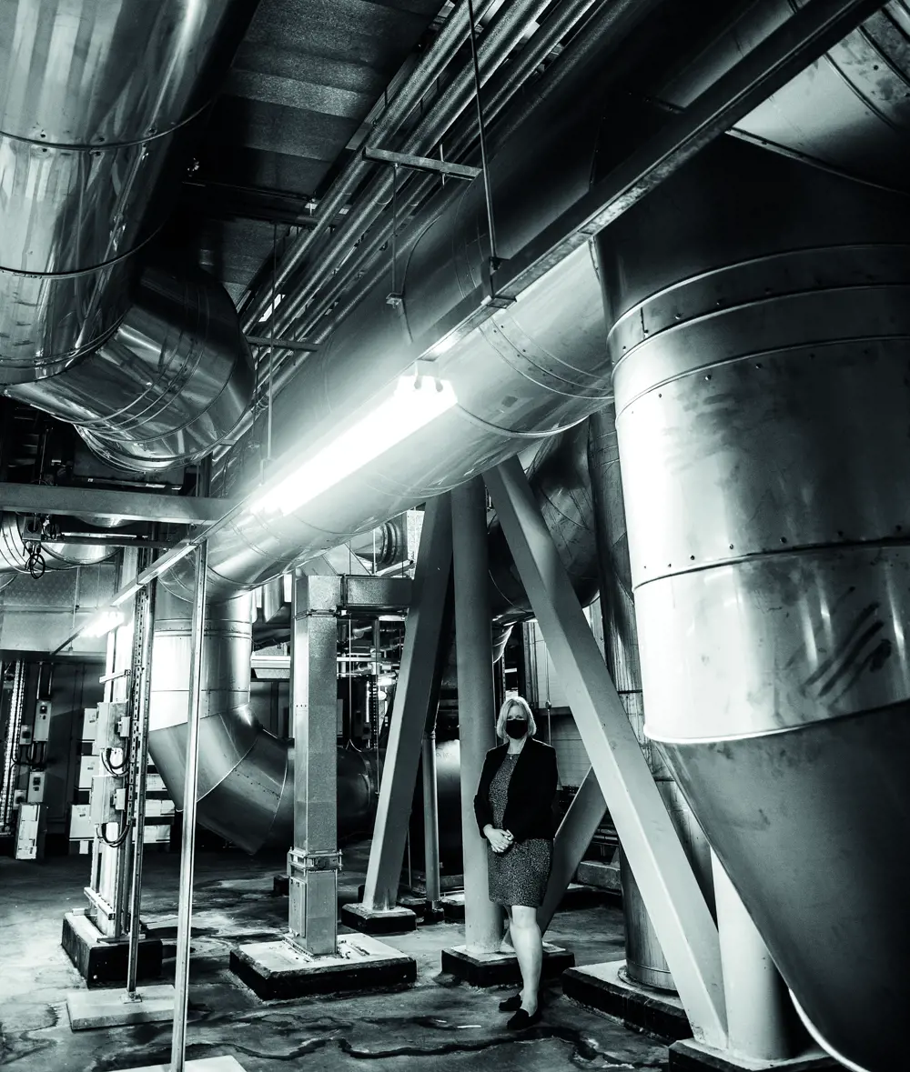 A black and white photo shows a blonde woman wearing a face mask. She is standing among some large ventilation tubes