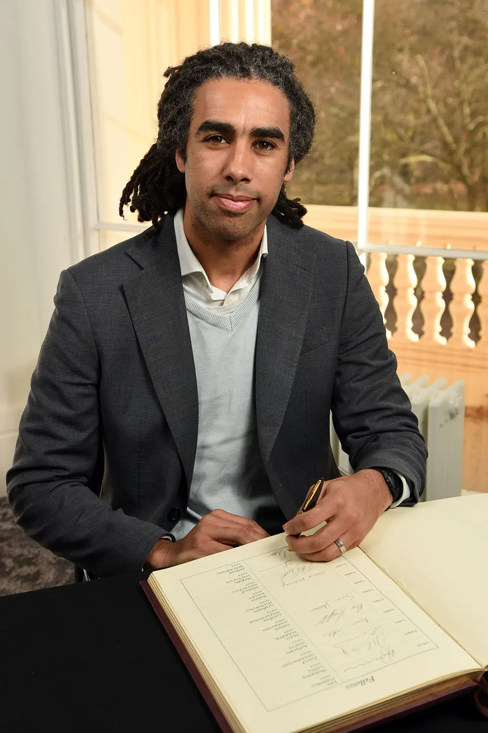 A person in a suit sits at a table and signs a large open book with handwritten entries, with a bright window and balustrade in the background.