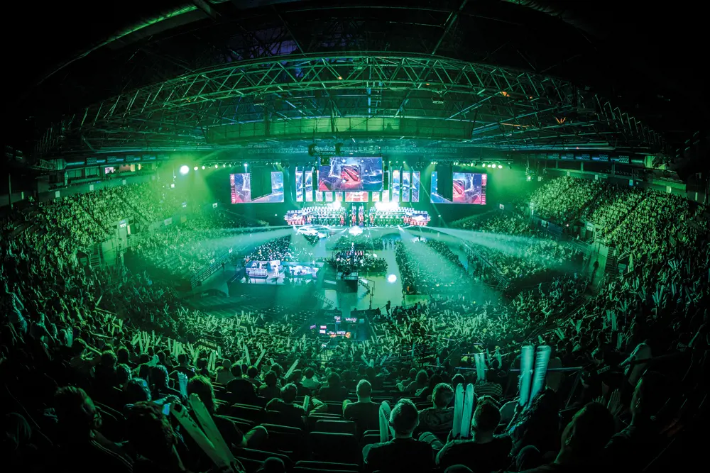 A crowd in a stadium watching a livestream of a gaming tournament in Birmingham with lights illuminating the audience.